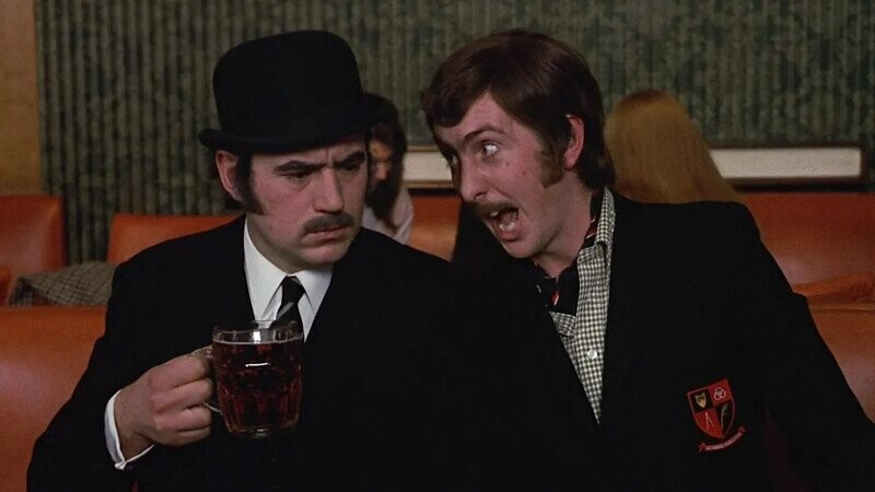 Two men in a pub: one in a bowler hat holds a pint while the other leans in, eyebrows raised, to nudge him.